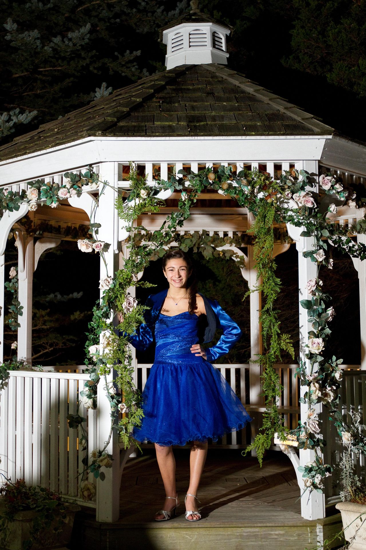 Bat mitzvah girl in a royal blue gown posing in a flower-draped gazebo at night