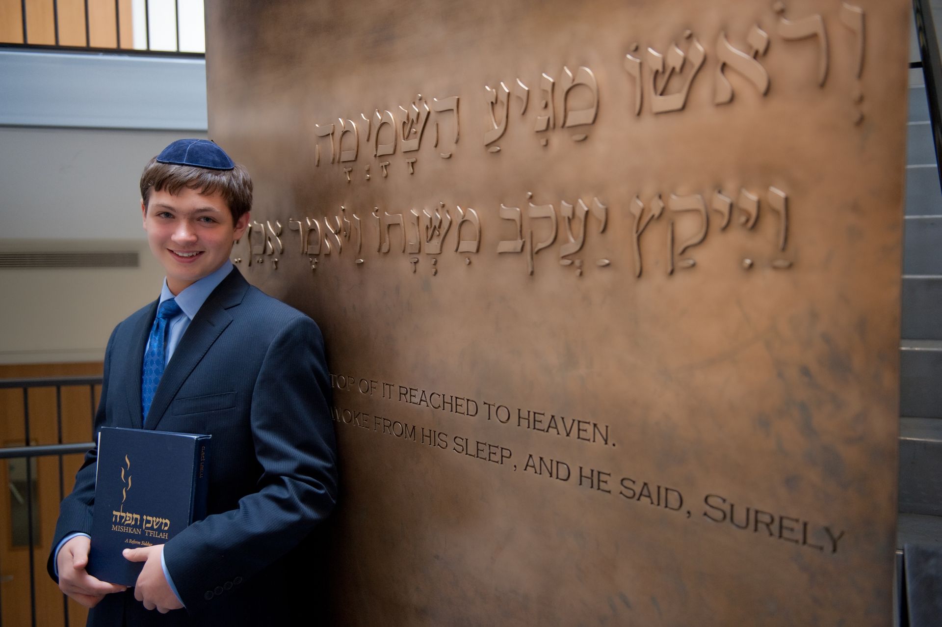 Bar mitzvah boy in a blue kippah and suit holding a siddur beside a bronze synagogue wall with Hebrew text