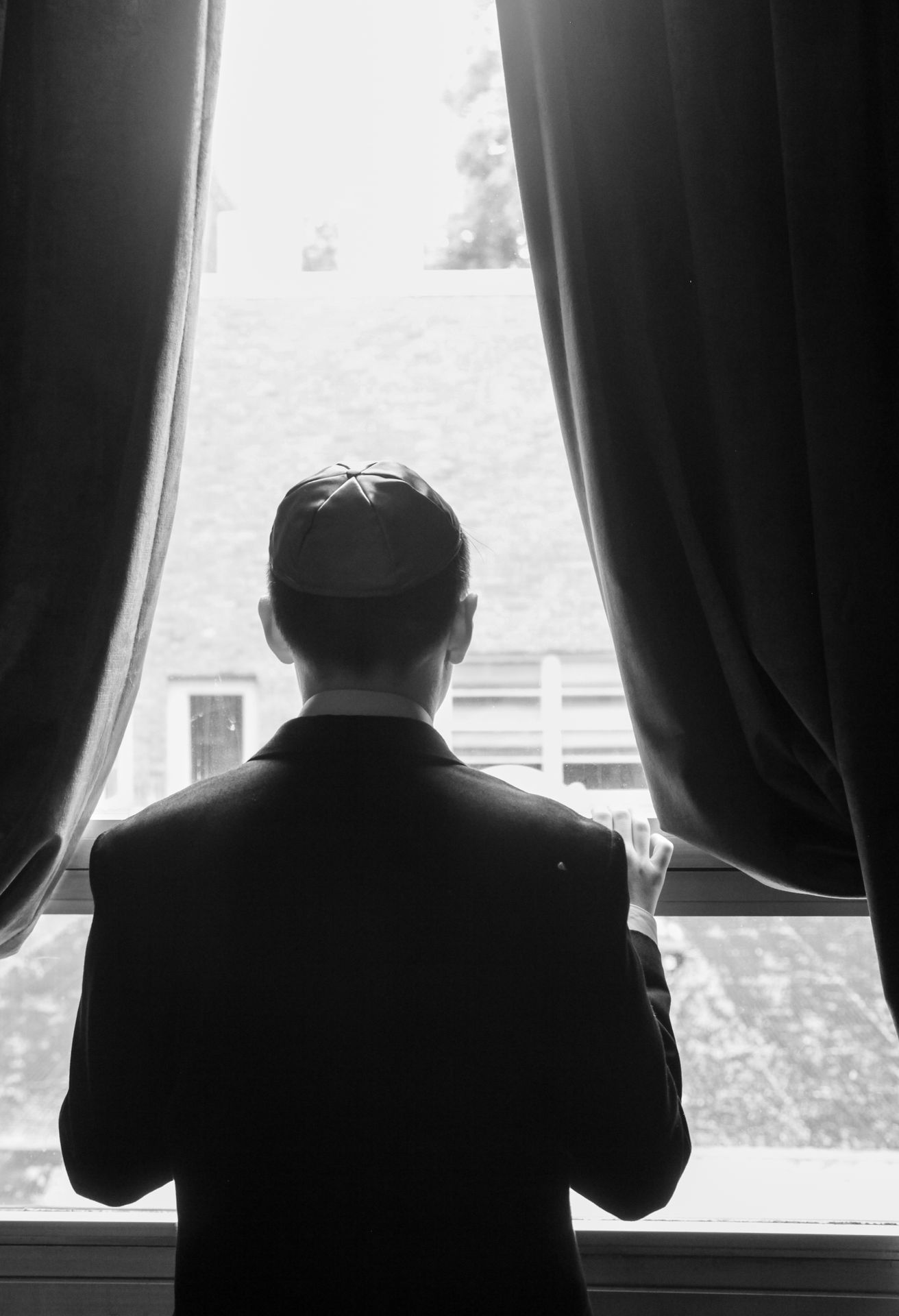 Black-and-white silhouette of a bar mitzvah boy in a kippah framed between dark curtains at a tall window