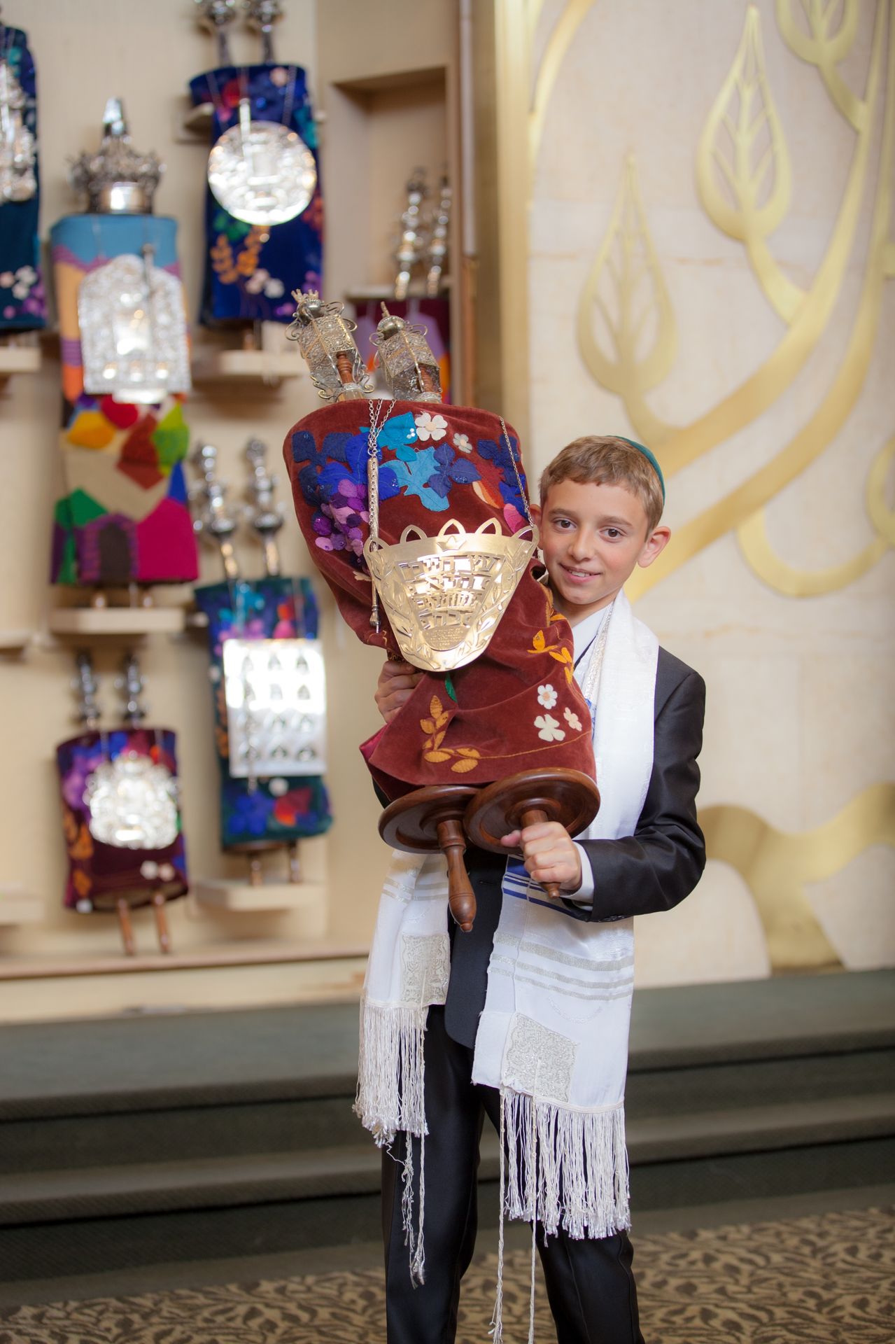 Bar mitzvah boy in a kippah and tallit holding a decorated Torah scroll before an ark of Torah mantles