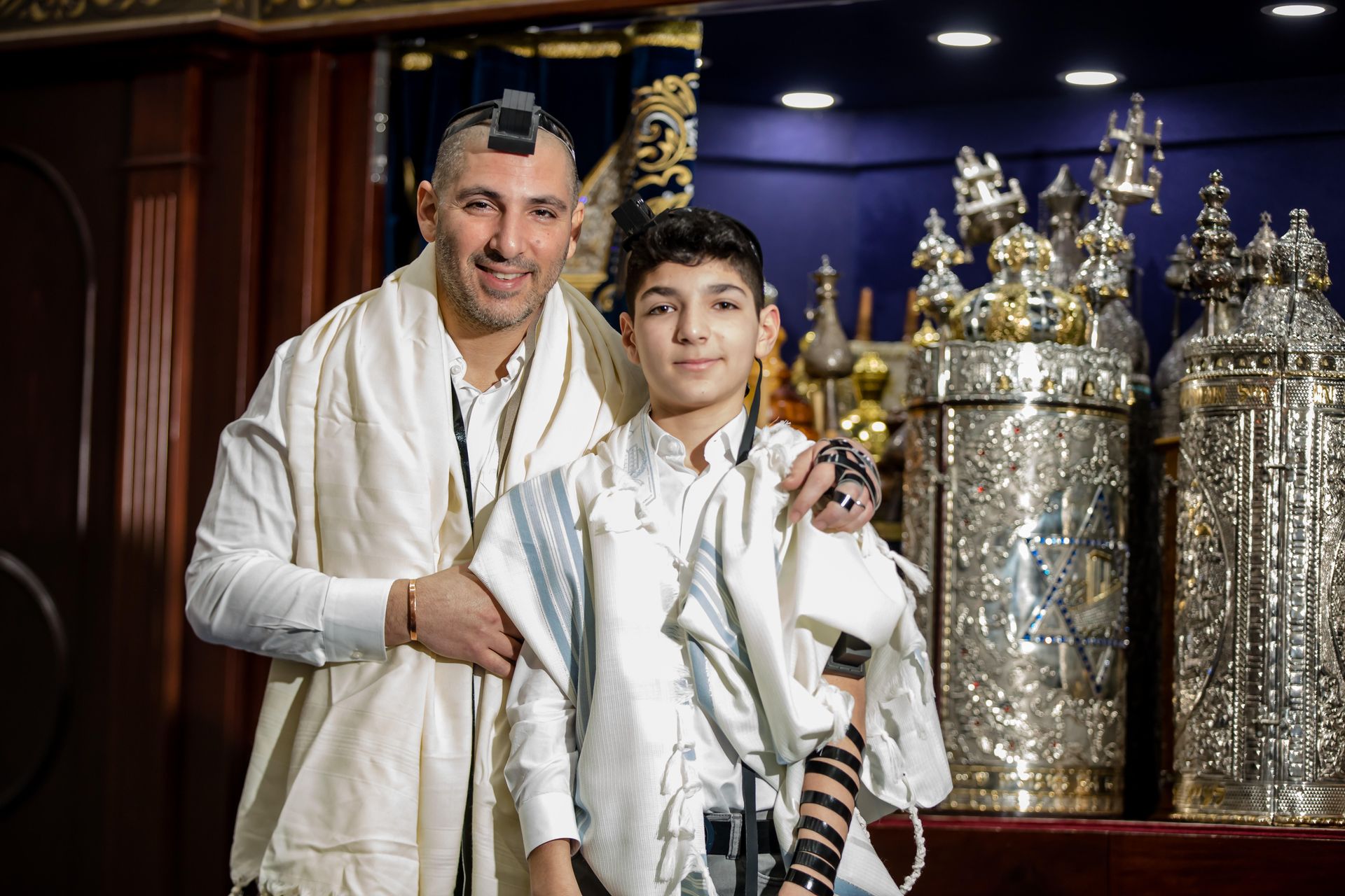 Bar mitzvah boy in a white tallit and tefillin posed with his father in a sanctuary lined with Torah crowns