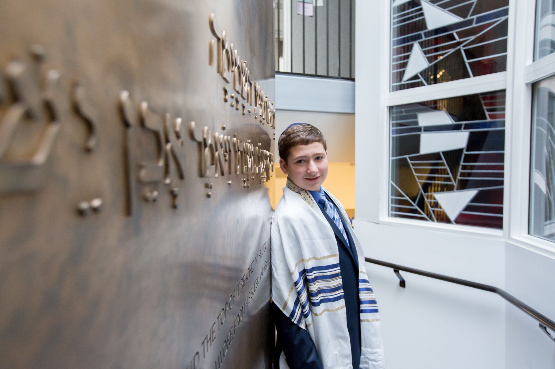 Bar mitzvah boy in a tallit leaning against an engraved Hebrew wall beside a patterned window