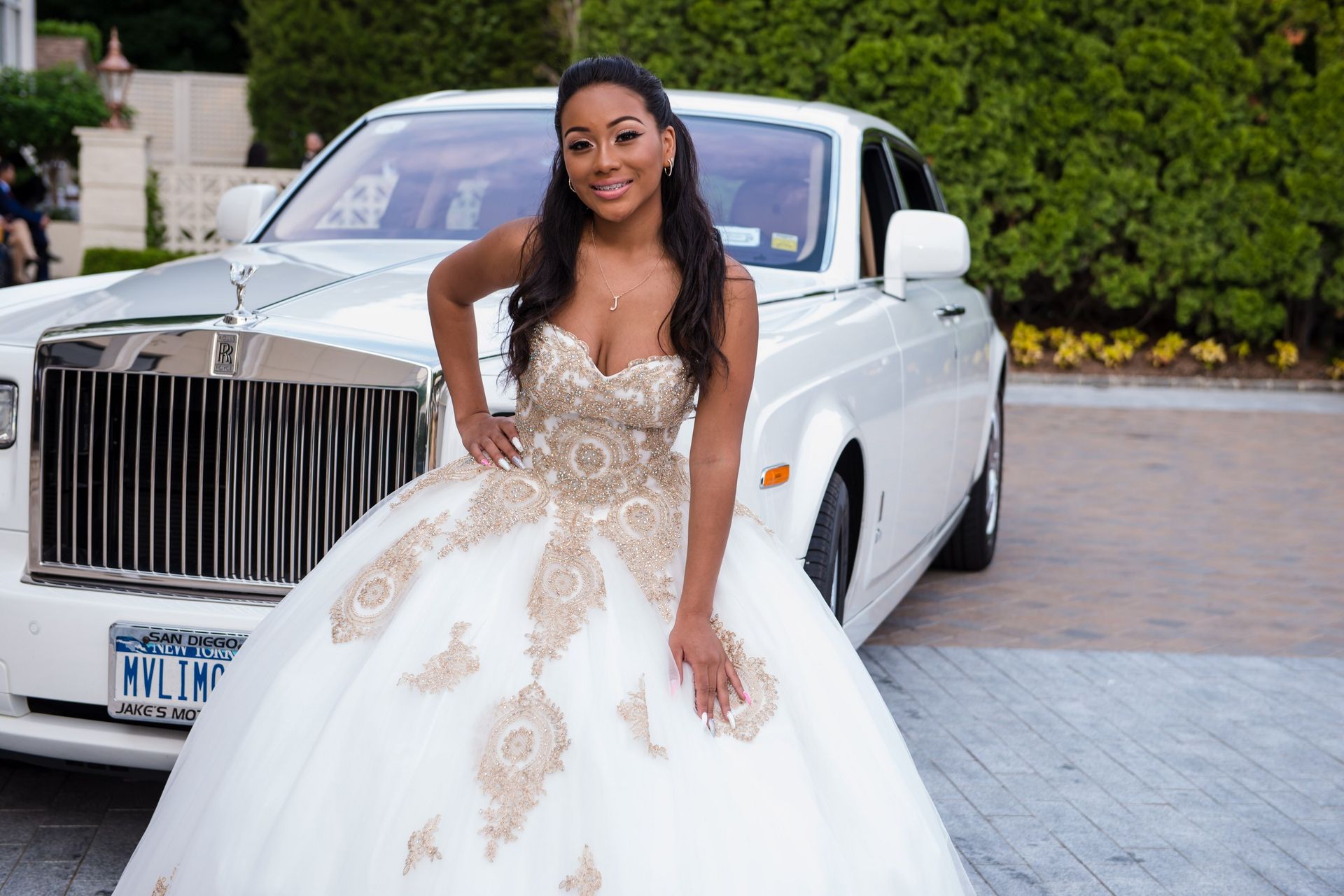 Quinceañera in a white and gold gown posing beside a white Rolls-Royce