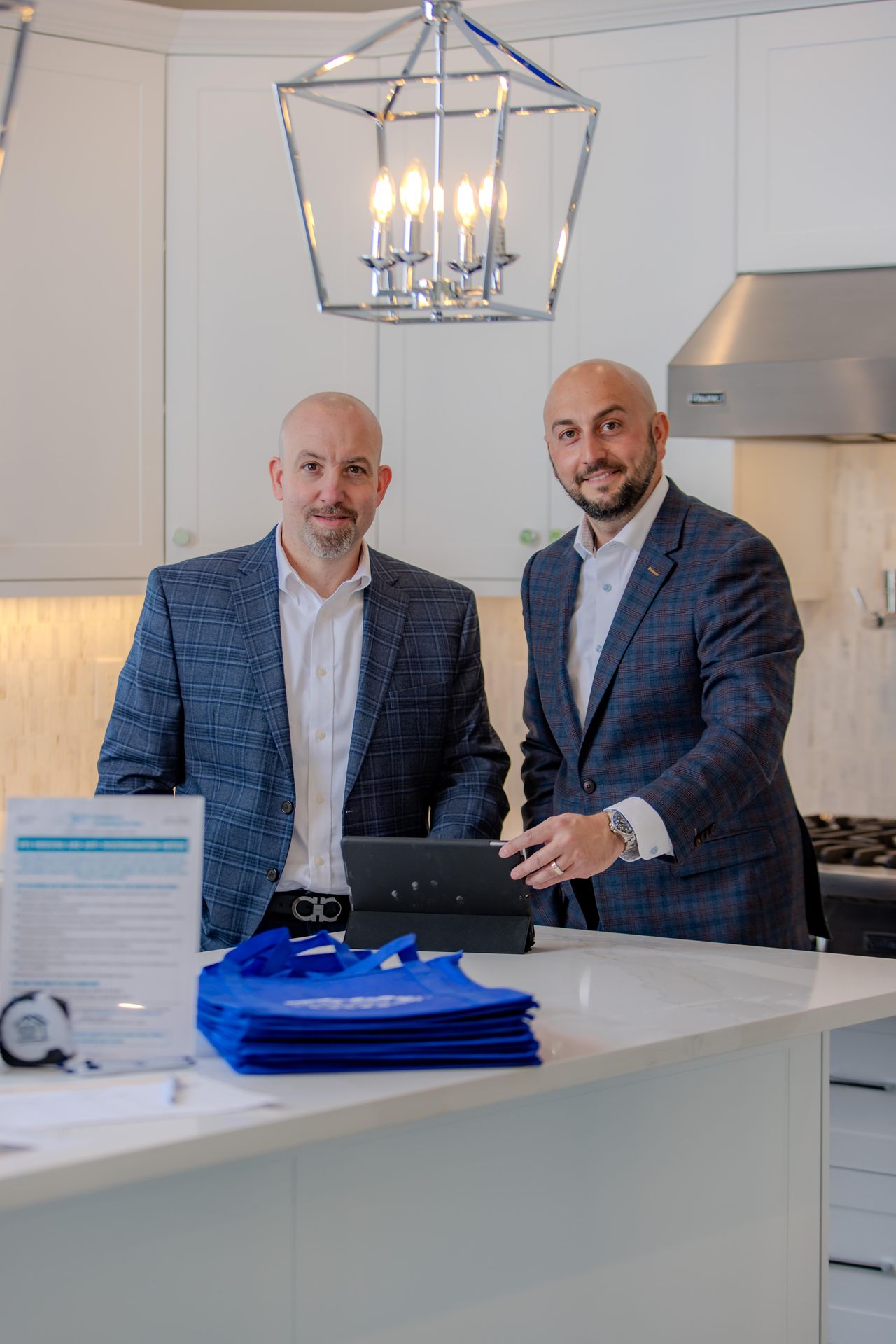 Two bald men in matching blue plaid blazers at a kitchen island with a tablet and blue bags